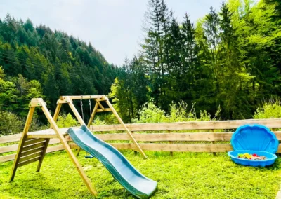 Holzspielplatz mit Rutsche, Kletterelement und Sandmuschel im Grünen.