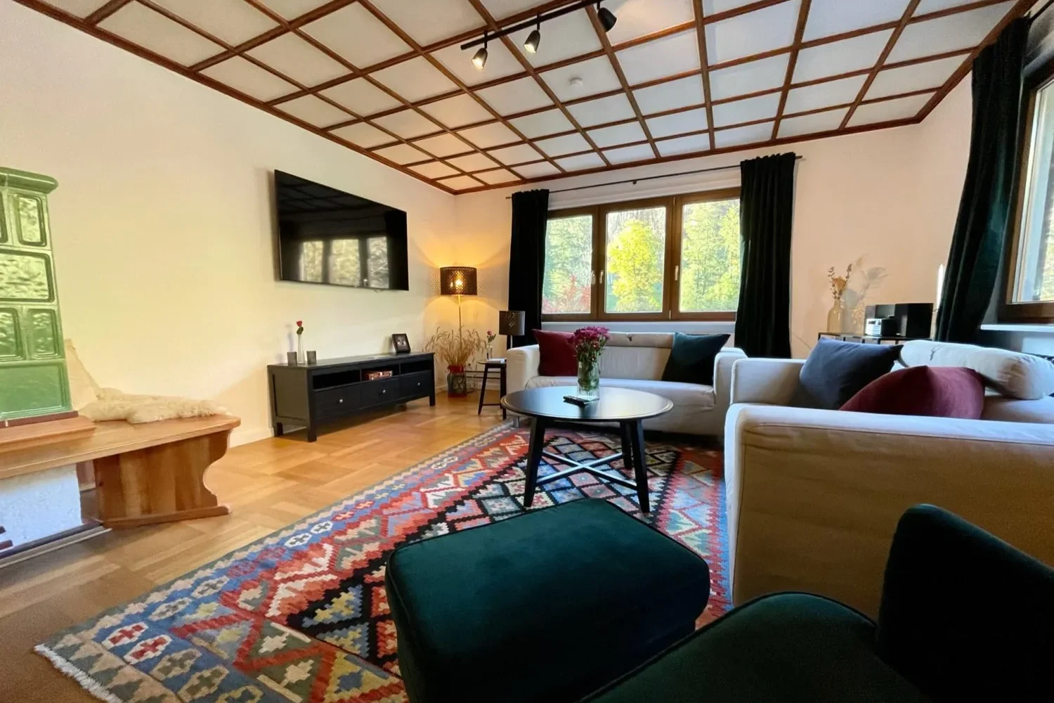 Wohnzimmer der Gruppenunterkunft Schwarzwald – Gemütlicher Sitzbereich mit Kachelofen Gemütliches Wohnzimmer mit zwei Sofas, buntem Teppich, Kachelofen und großem Fenster in der Gruppenunterkunft Schwarzwald.