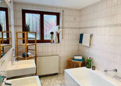 Modernes Badezimmer der Ferienwohnung Bergsee mit Badewanne, zwei Waschbecken und heller Fliesenwand.