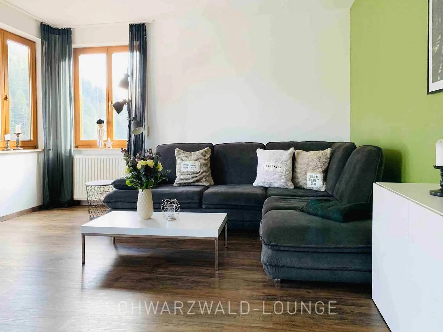 Wohnzimmeransicht im Apartment Bergsee Schwarzwald mit großer Sofalandschaft, Blumen und heller Fensterfront.